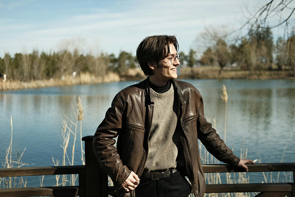 Man in a Brown Leather Jacket and Gray Sweater on a Footbridge over a River