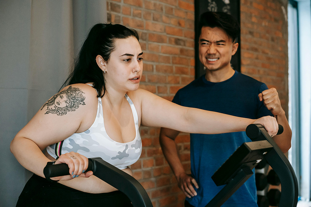 Young woman training hard on elliptical machine with support of trainer