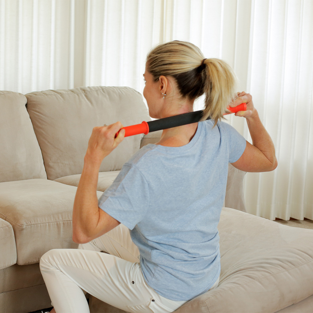 A woman sits on the couch using Tiger Tail Original 18 handheld massage roller across her upper back and neck