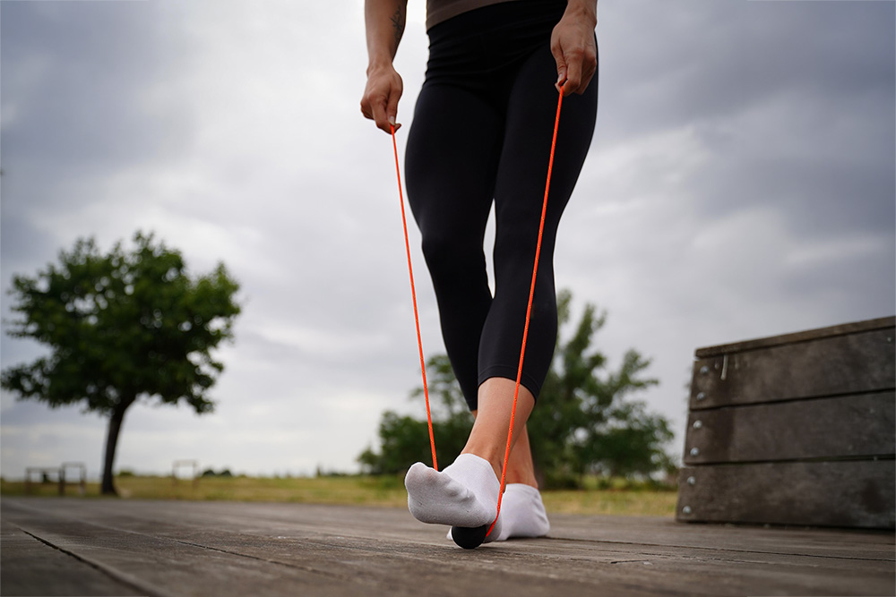 A person rolling a firm massage ball under the sole of their foot from heel to toe while standing.